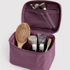 A purple zippered travel or toiletry bag shown open, containing a wooden hairbrush, small bottles, a tube, and makeup brushes, on a light gray background.